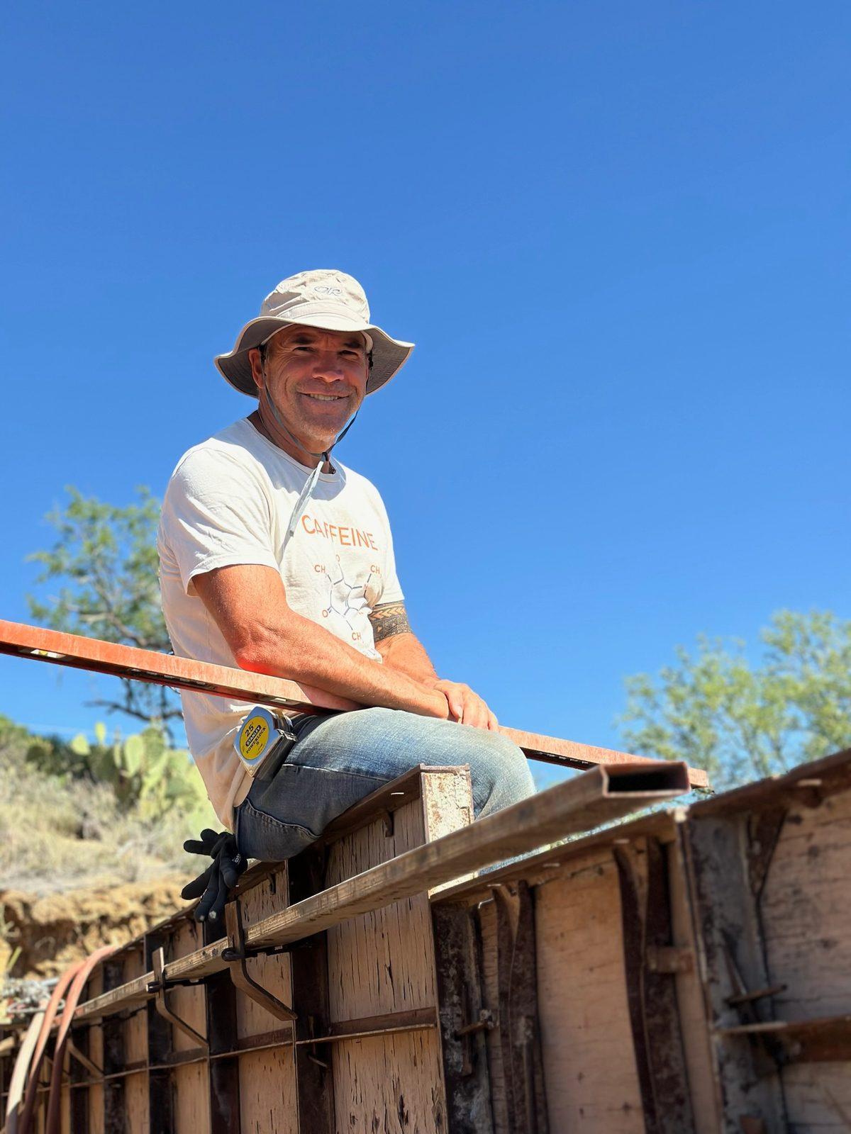 Brian Johnson sitting on top of the rammed-earth wall forms in a sun hat