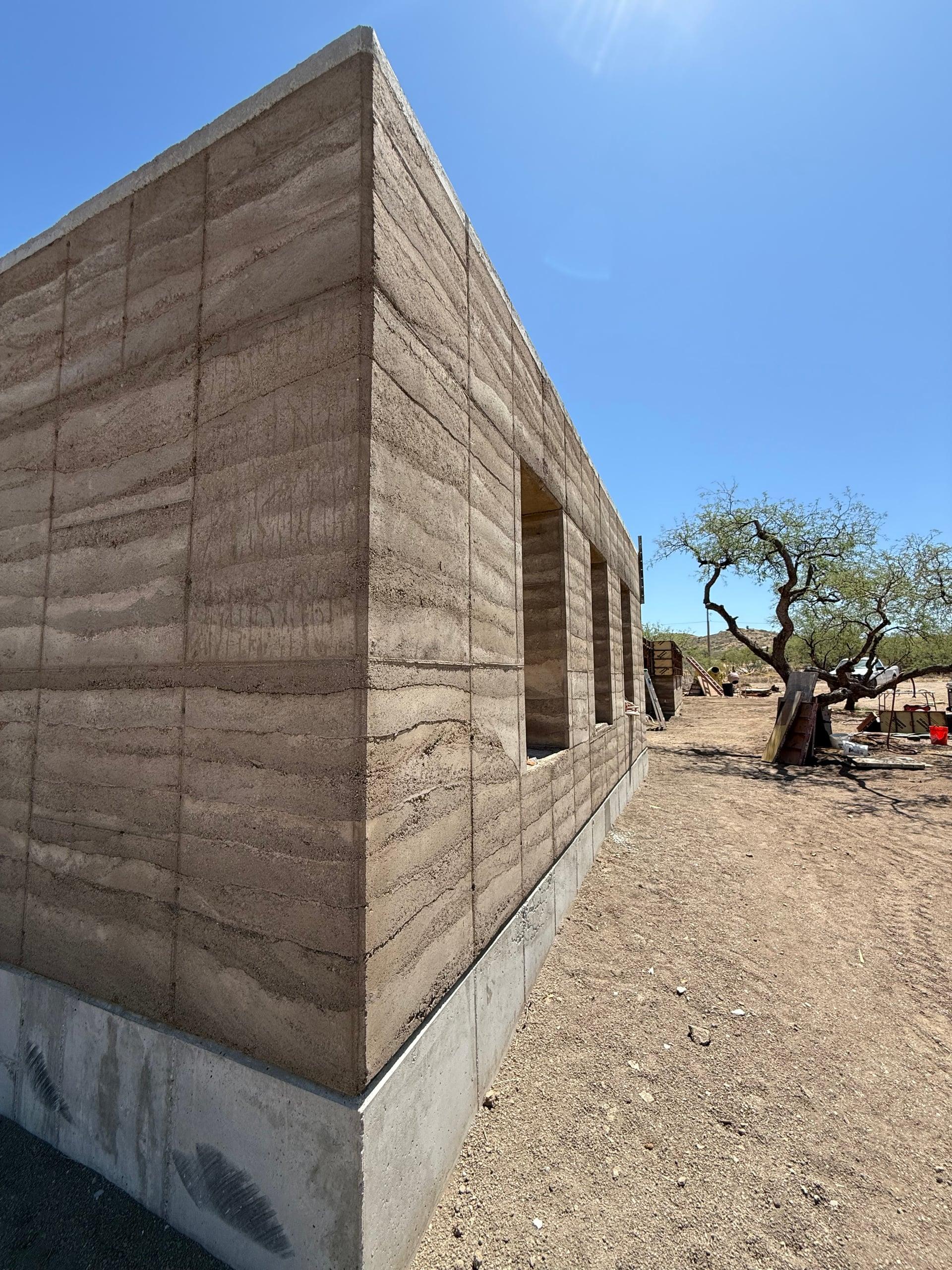 Cured rammed-earth corner with window opening beside a mesquite tree