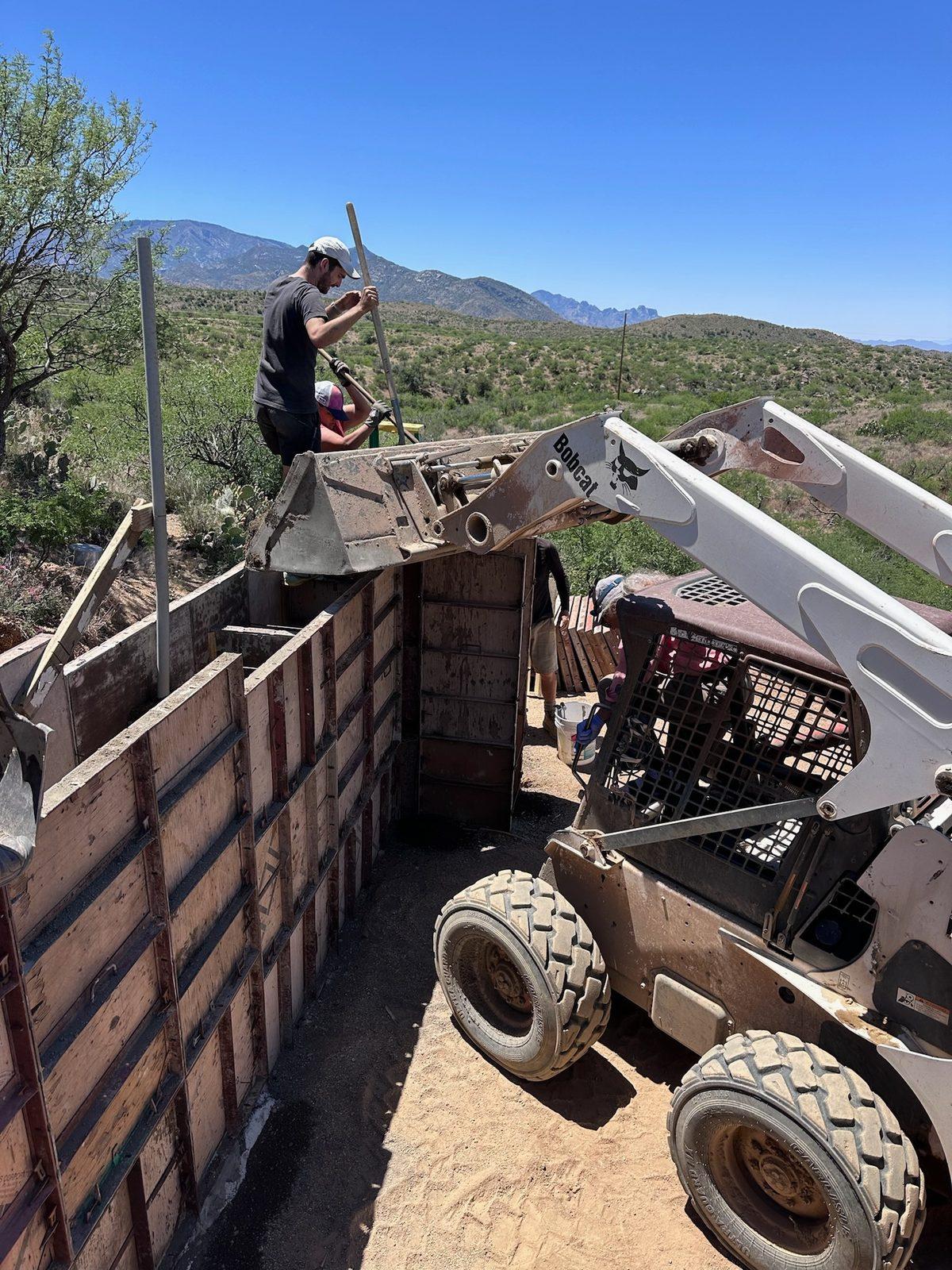 Skid steer loading soil into rammed-earth wall forms while a crew member works on top of the forms