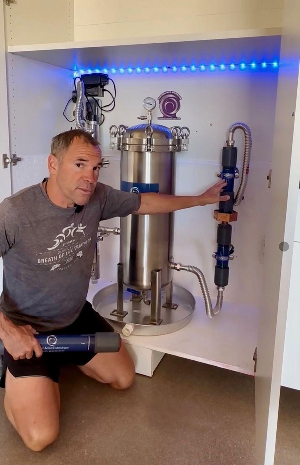 Brian Johnson kneeling beside the stainless structured water vessel in its lit cabinet
