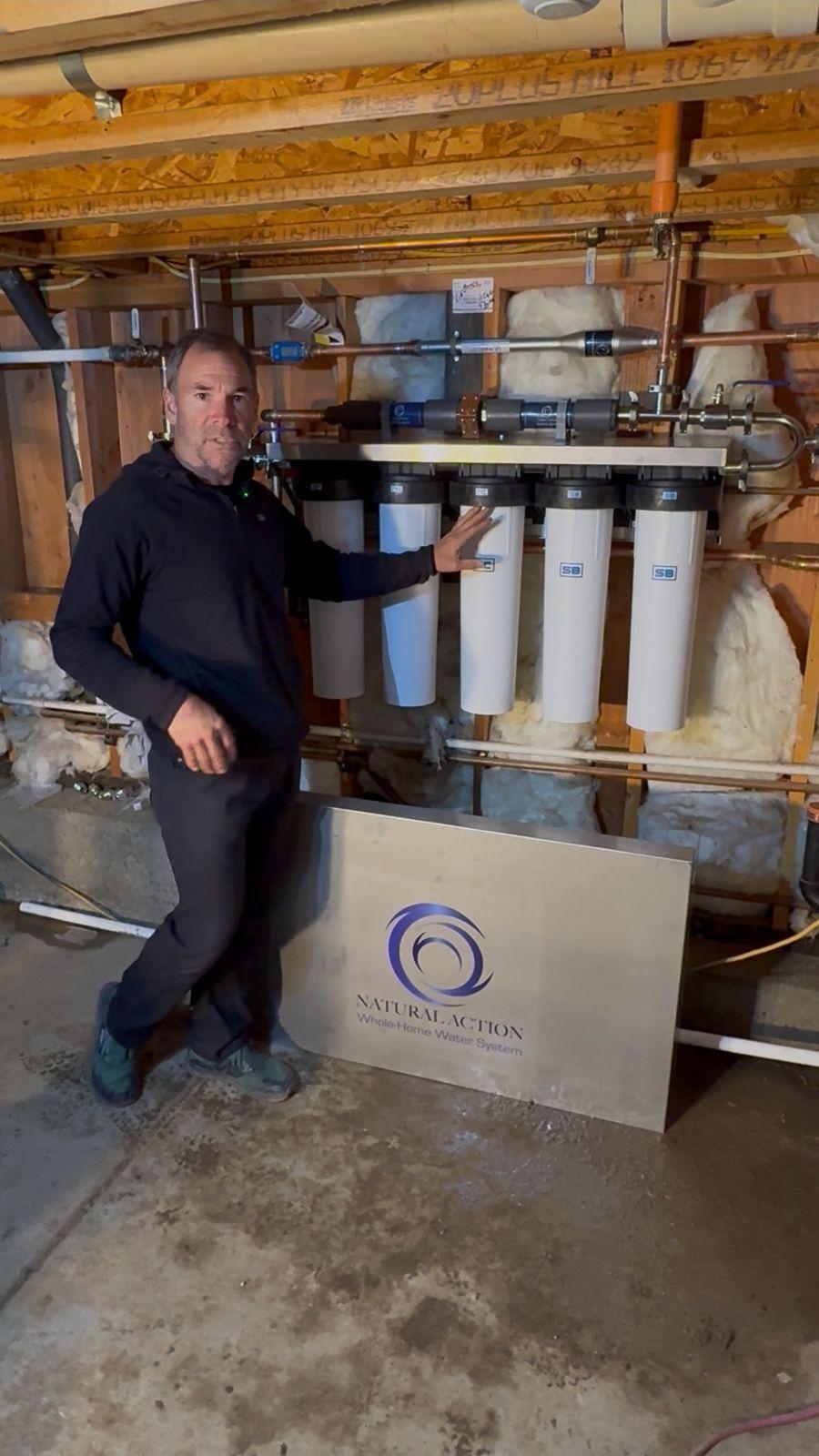 Brian Johnson beside a five-cartridge whole-home water system in a basement, with the stainless enclosure cover leaning below