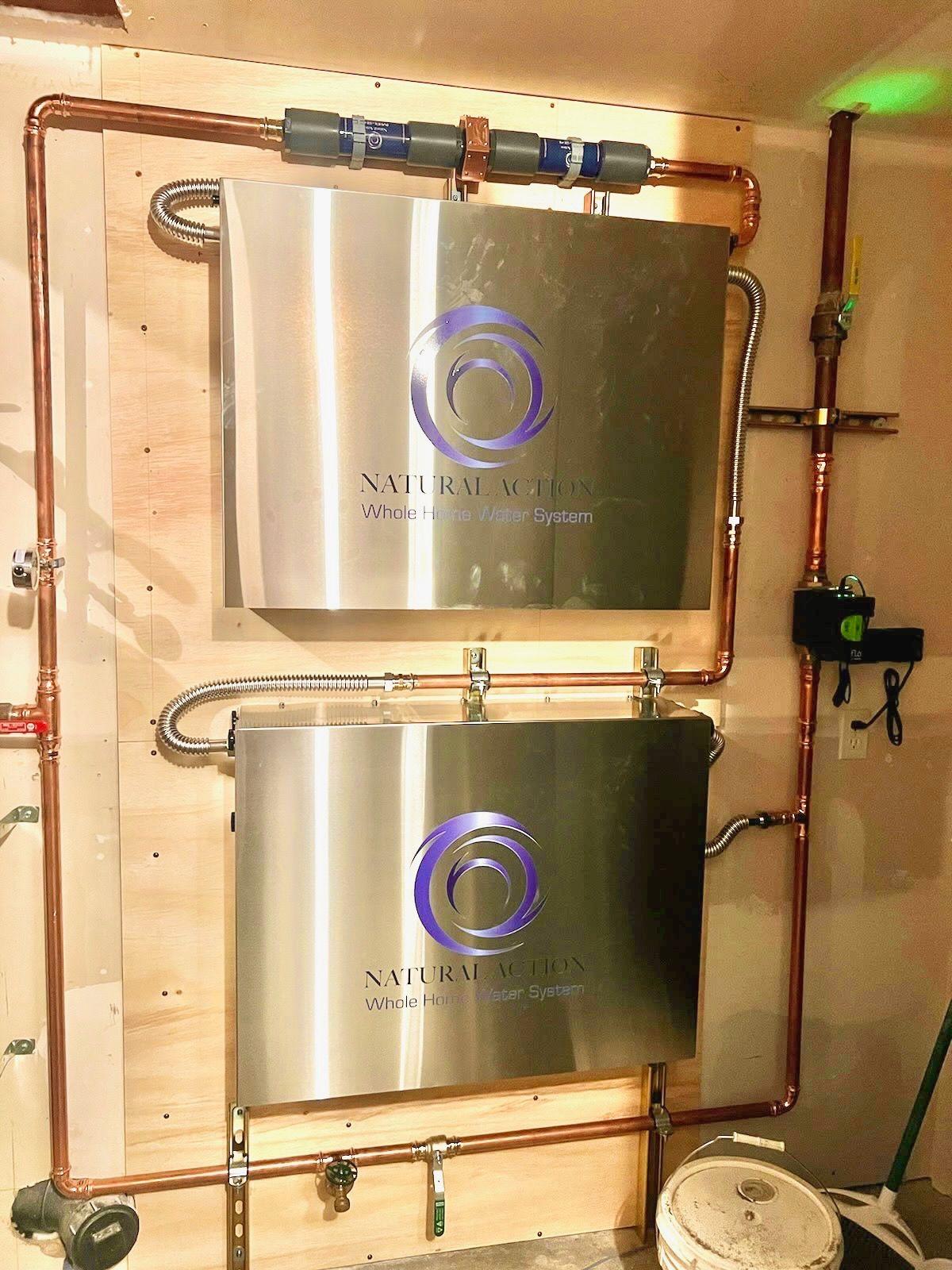 Two stainless Natural Action enclosures mounted one above the other on a plywood wall with copper piping