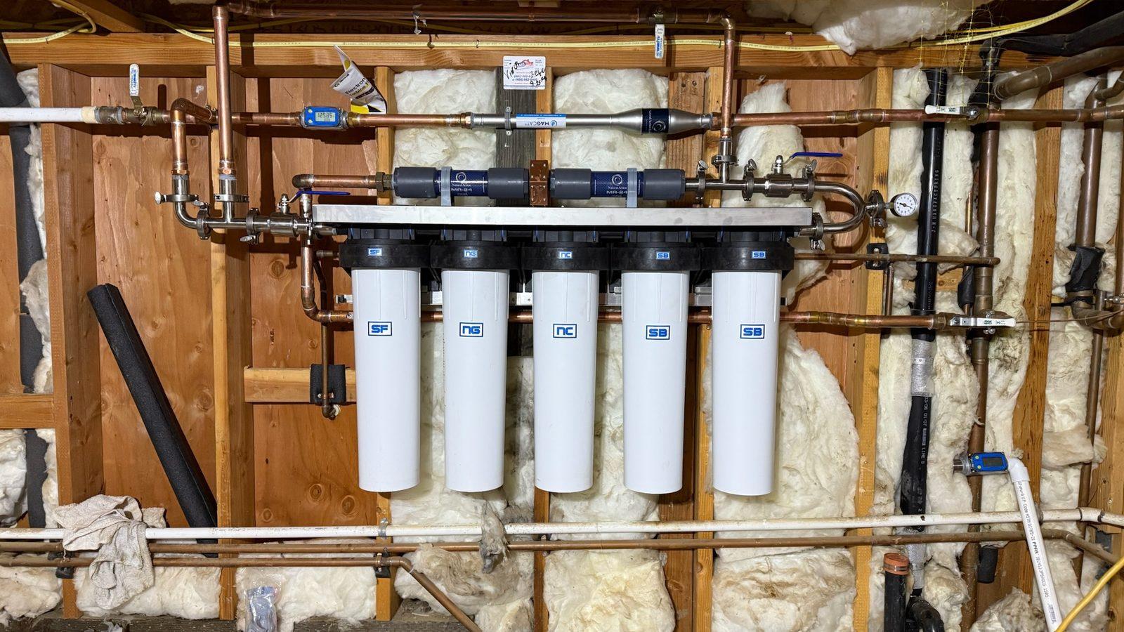 Whole-home water treatment wall roughed in on open framing: five cartridge housings under a manifold of inline structured-water units and copper piping