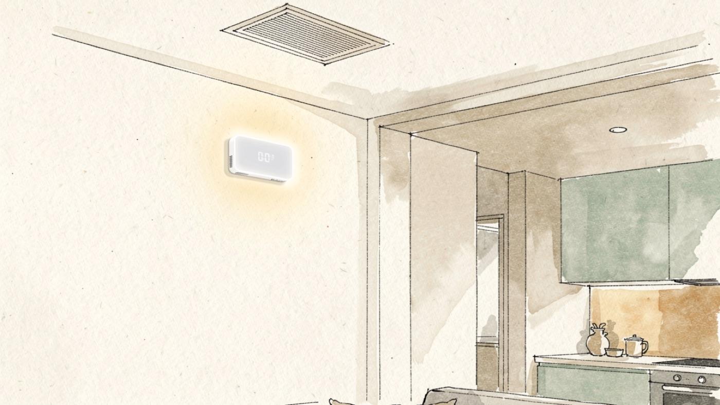 Living room wall with a small air quality monitor mounted at chest height, its display glowing, near an open kitchen and a ventilation grille